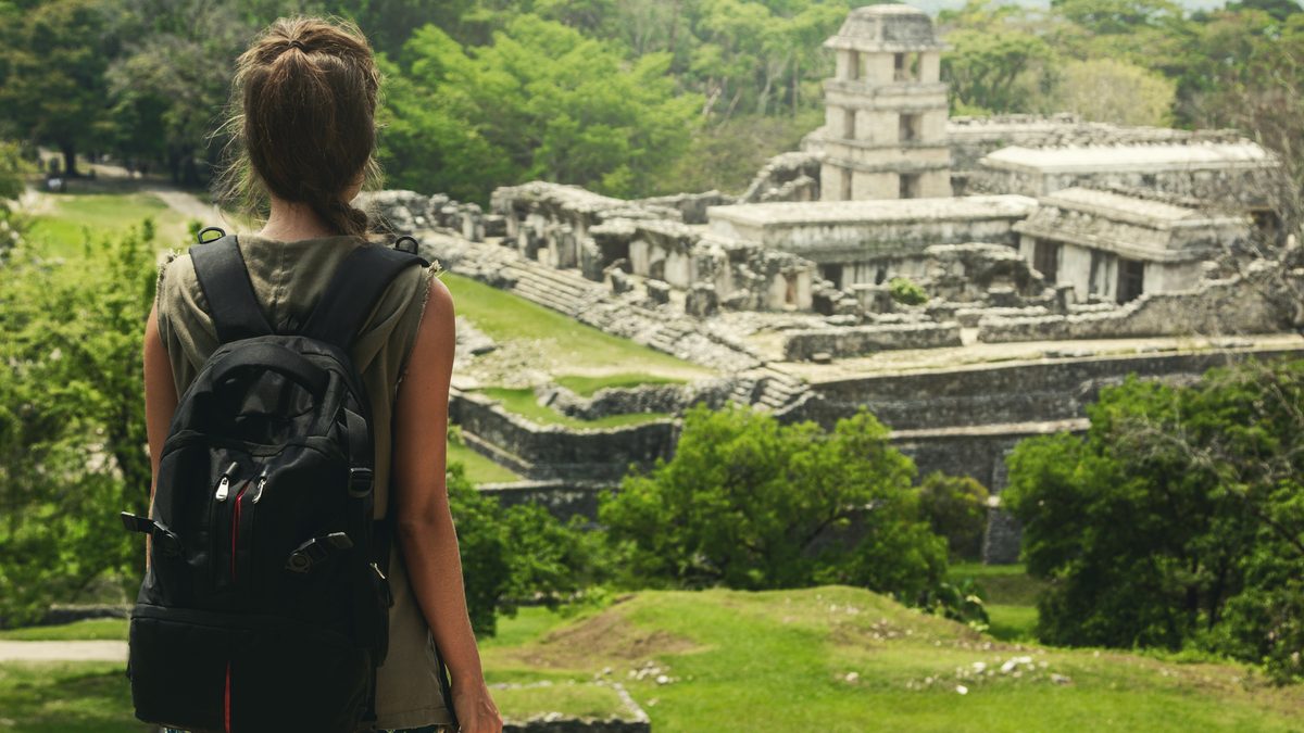 A backpacker seen from behind looking over stone Mayan ruins surrounded by forest.